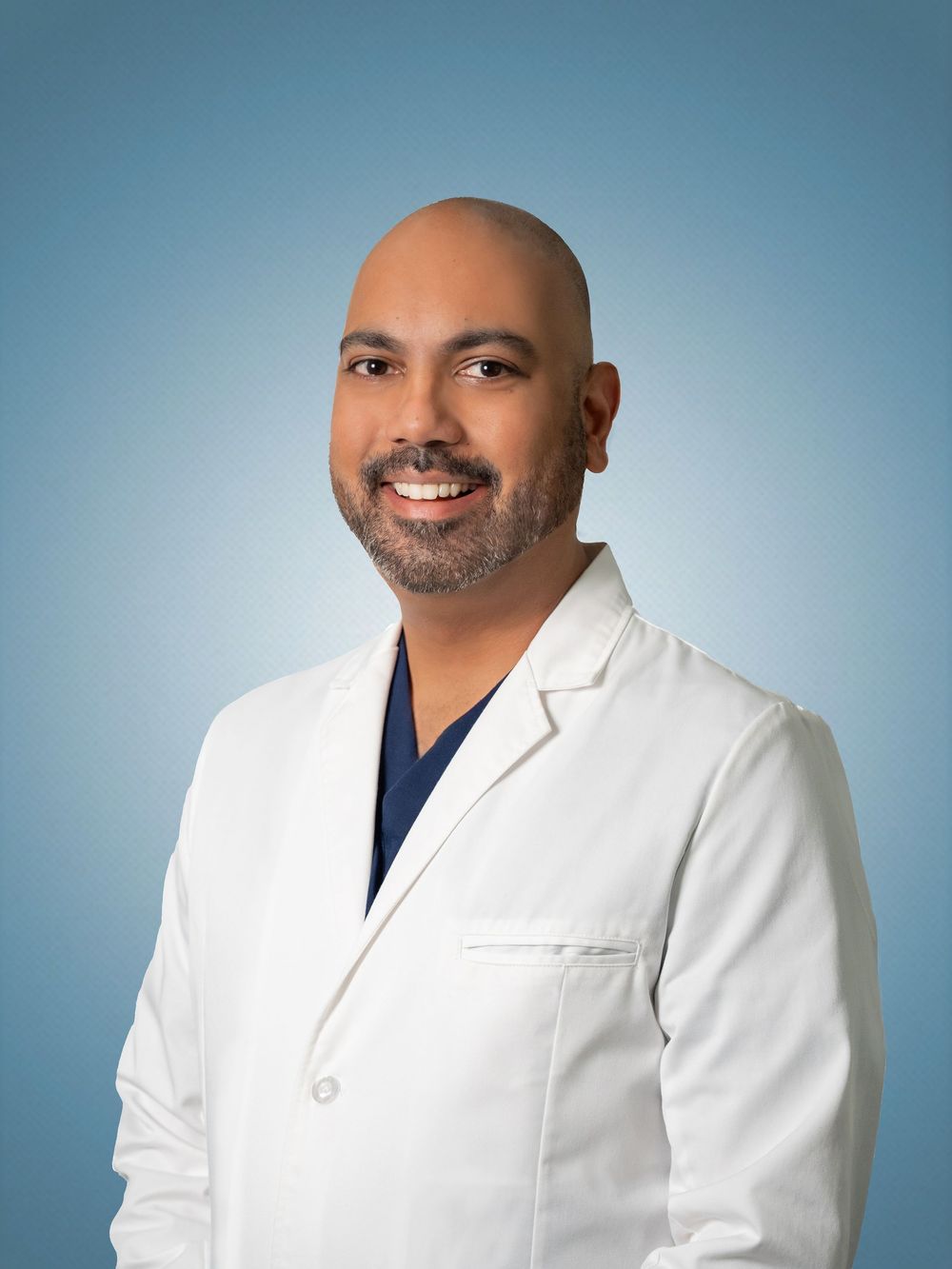 Professional headshot portrait of a medical practitioner wearing a white lab coat and navy scrub top against a blue background.