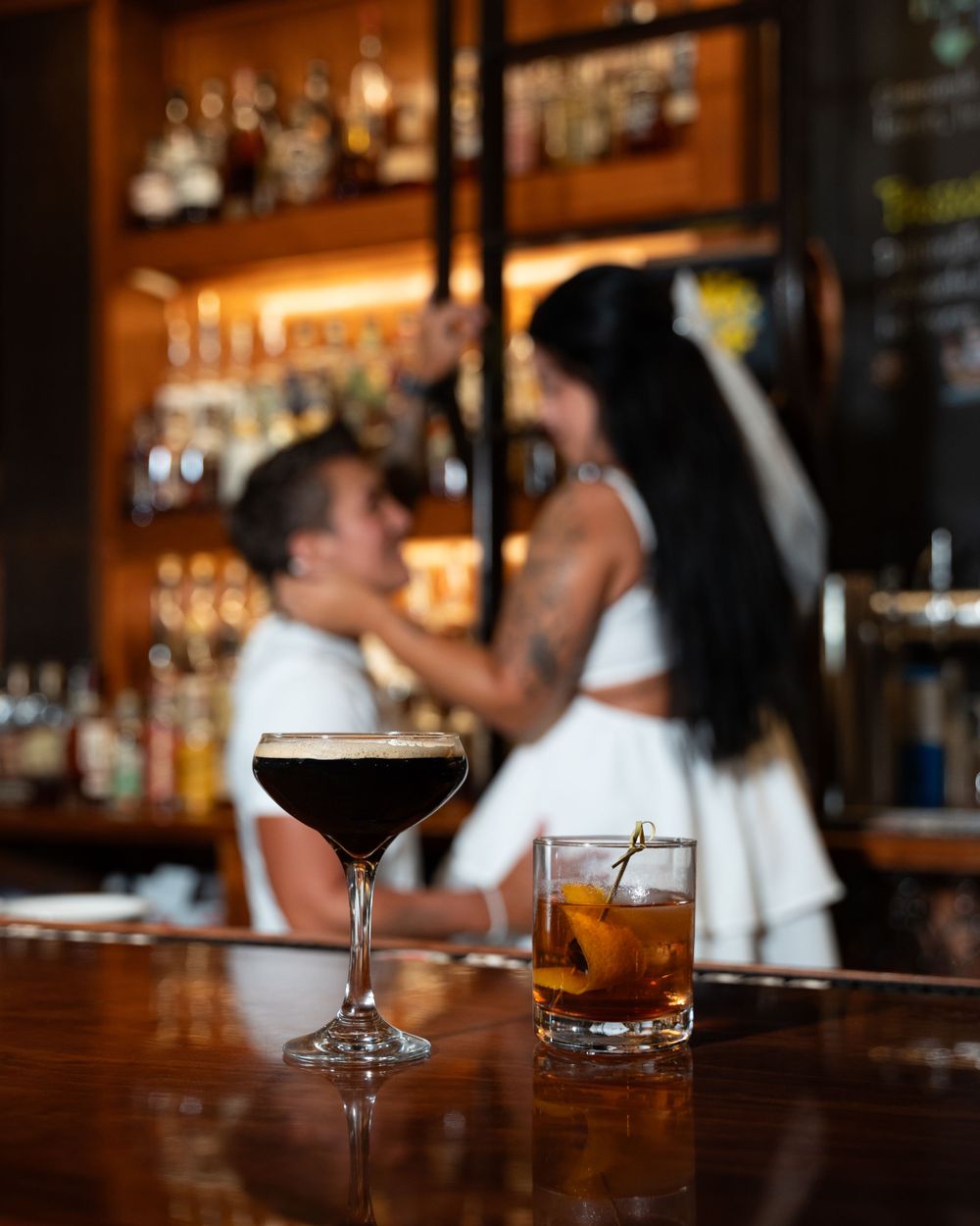 Two cocktails sit on a wooden bar counter in a dimly lit atmosphere with warm amber lighting, a couple embraces in the background.
