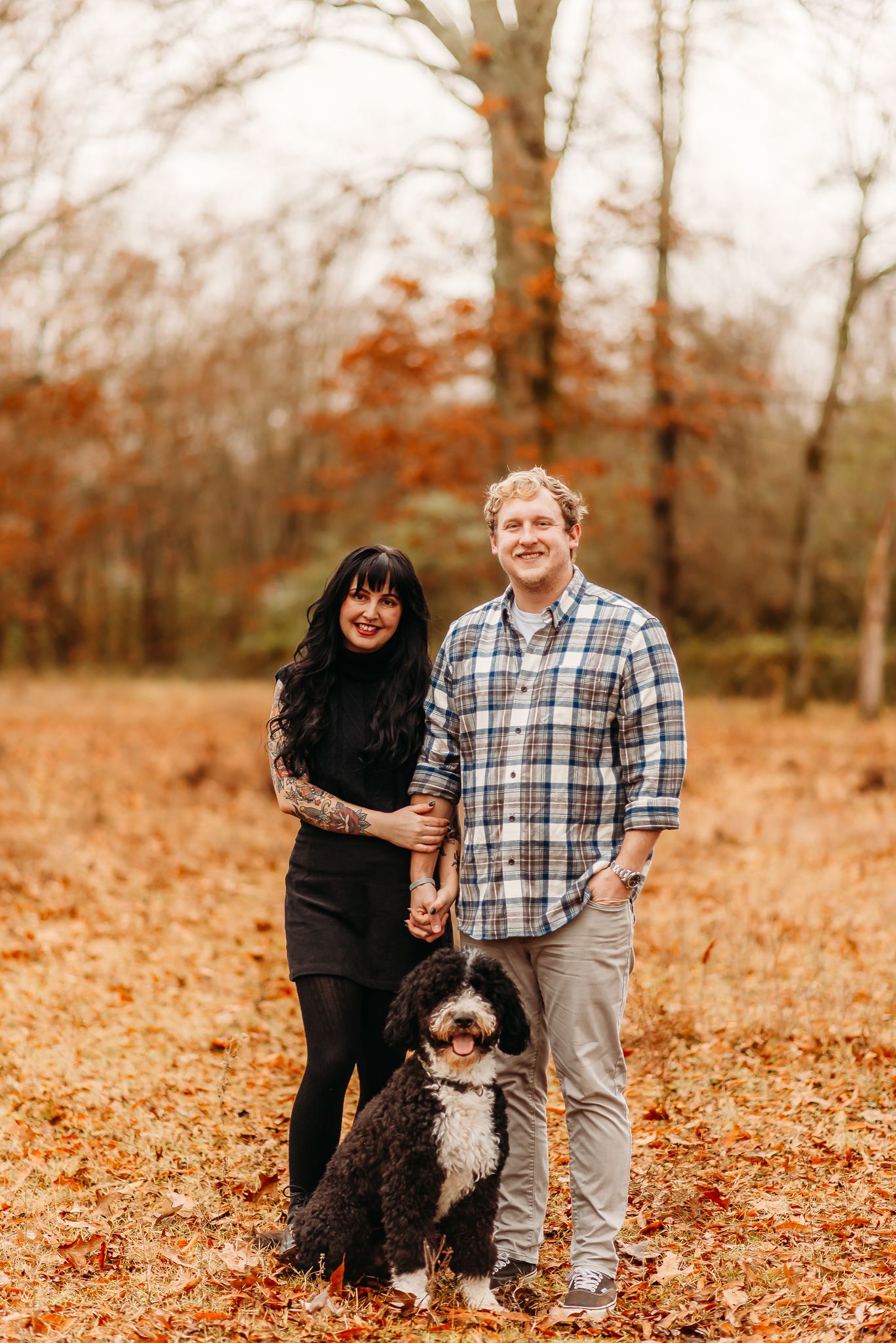 A couple and their Bernedoodle dog pose together on fallen autumn leaves in a wooded setting.