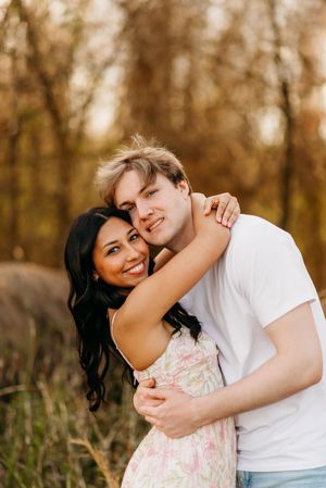 A couple embraces outdoors in a romantic pose during golden hour, surrounded by autumn trees and tall grass.