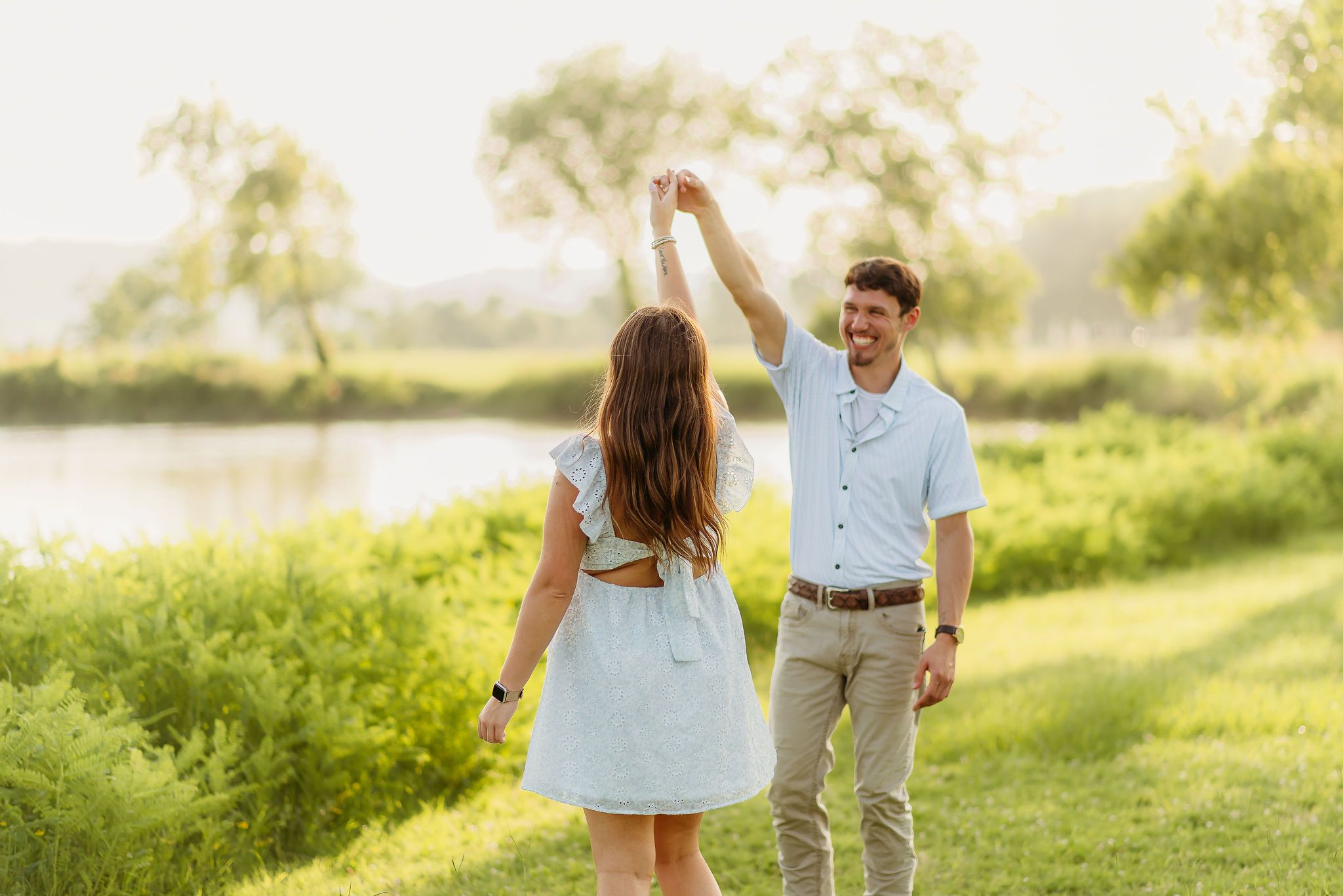 A couple dances together on a grassy hill overlooking a lake during a golden summer evening.