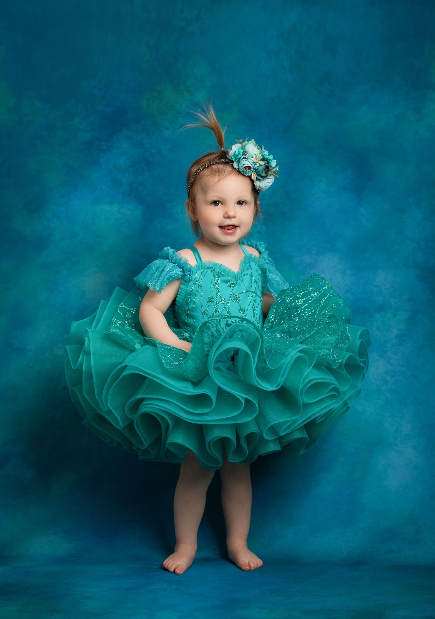 A baby wearing a ruffled turquoise dance costume and matching floral headpiece poses against a teal backdrop.