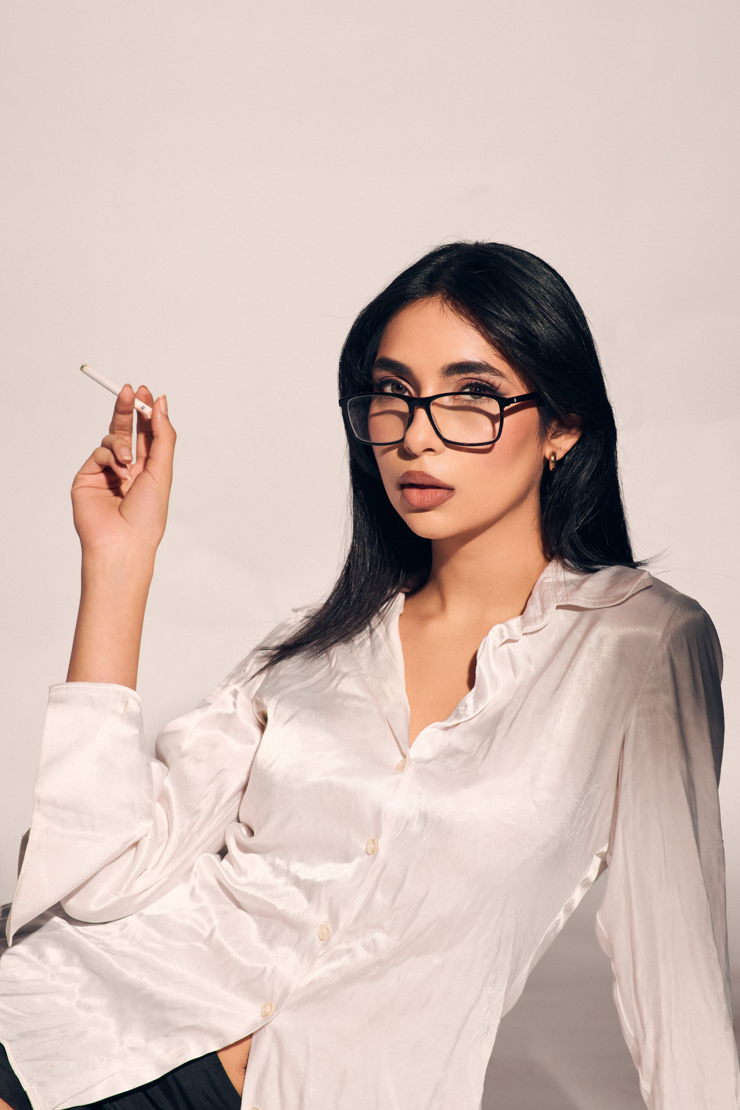 Woman in a white oversized shirt and black shorts, lying on her back with legs raised, posing stylishly against a neutral backdrop.