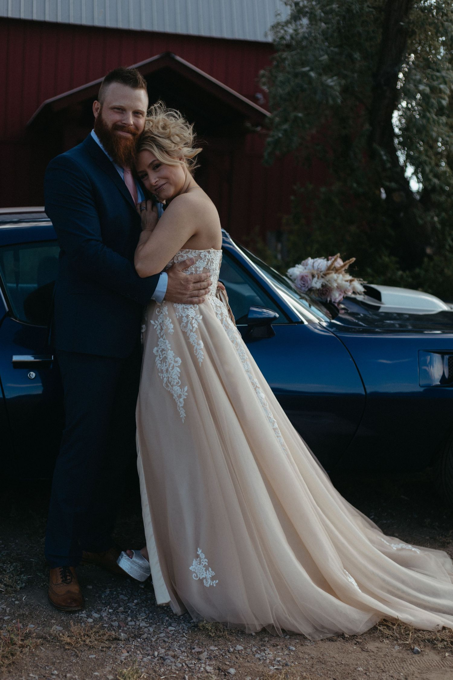 A bride in a champagne-colored lace gown poses near a vintage blue car and red barn.