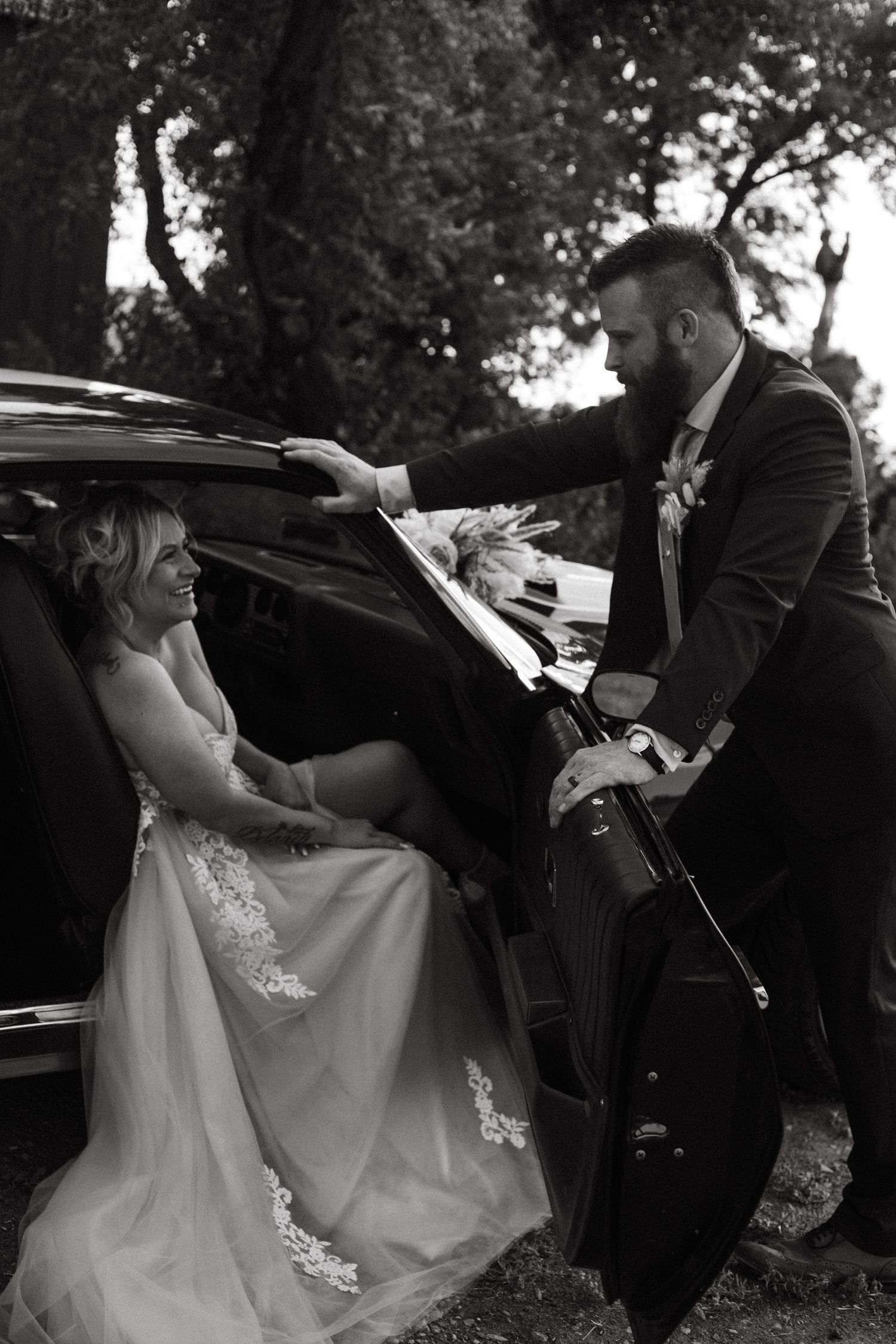 Black and white wedding photography capturing candid moments by a vintage automobile.