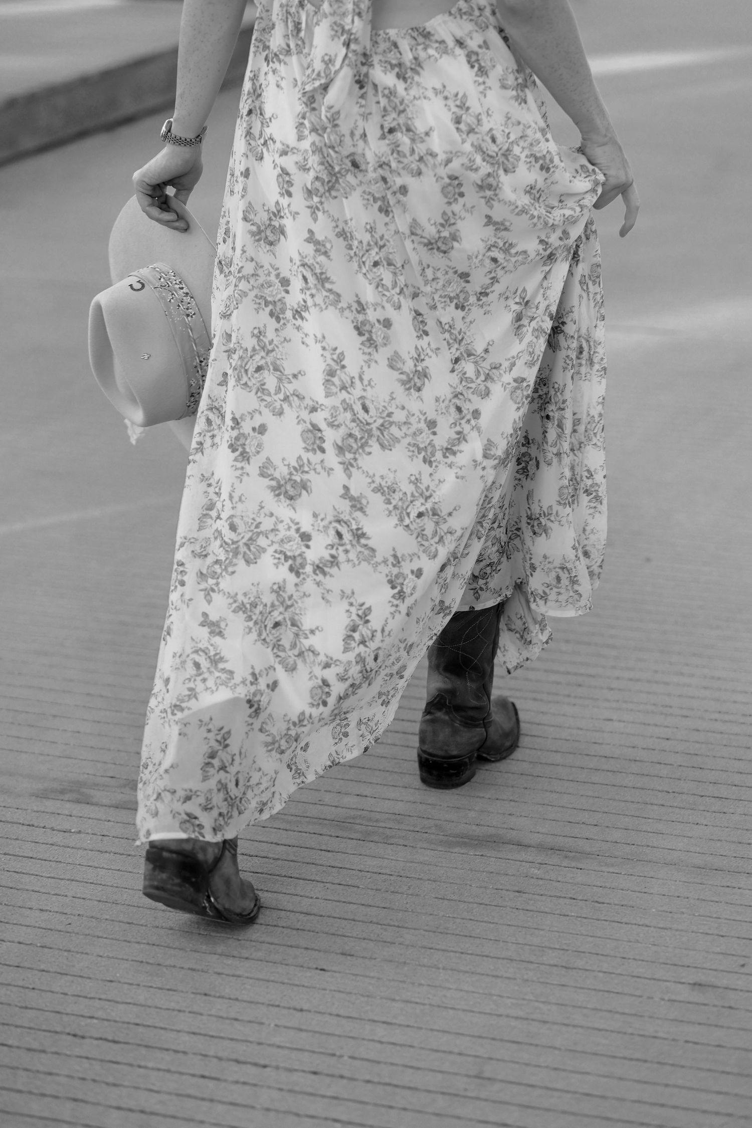 Black and white photo of a woman in a floral dress and boots walking while holding a hat.