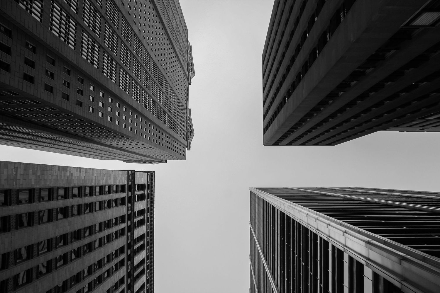 Looking up at modern skyscrapers converging to a point against cloudy sky in black and white.