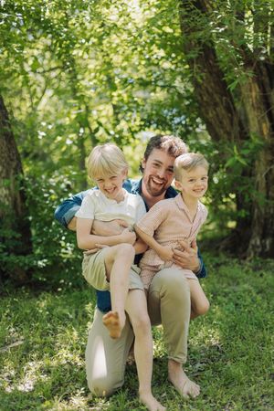 A smiling father kneels outdoors holding two young blonde boys in a lush green park setting.