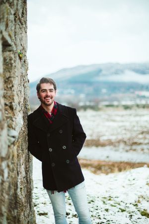 A person in a black peacoat stands against a snowy winter landscape with mountains in the background.