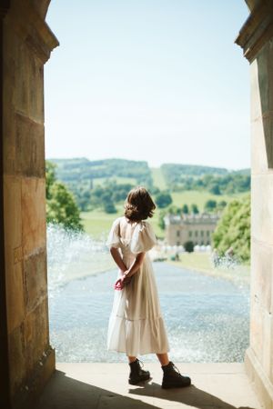 A person in a white dress looks out over scenic countryside from a stone archway.