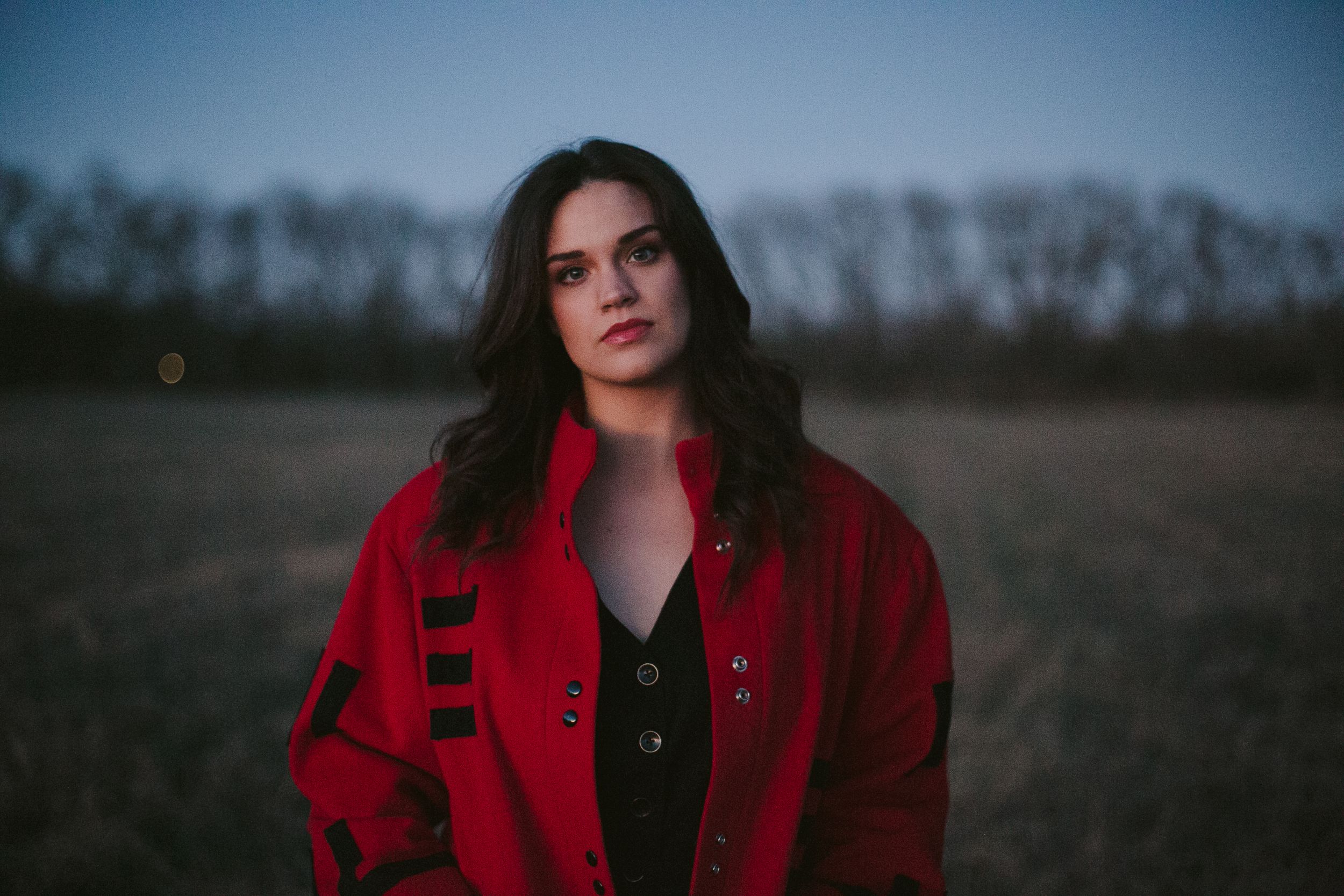 Person in a red coat poses outdoors at dusk against a rural backdrop.