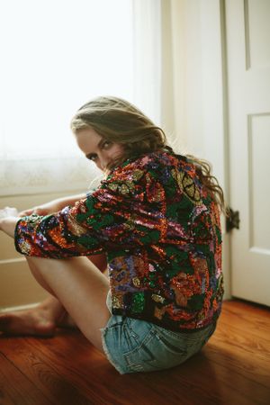 Person in floral print shirt and denim shorts sitting on hardwood floor near window.