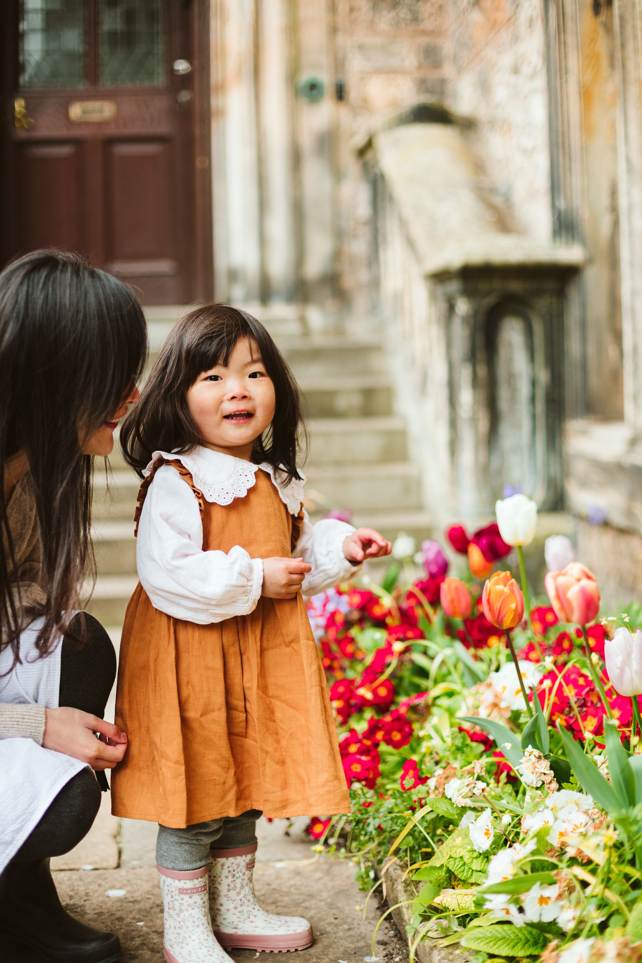 Smiling toddler girl in orange dress and white boots admiring colorful tulips with her mother outdoors.