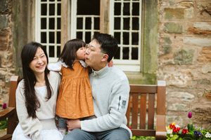 Happy Asian family with young daughter sharing a kiss outdoors on a bench near a stone building.