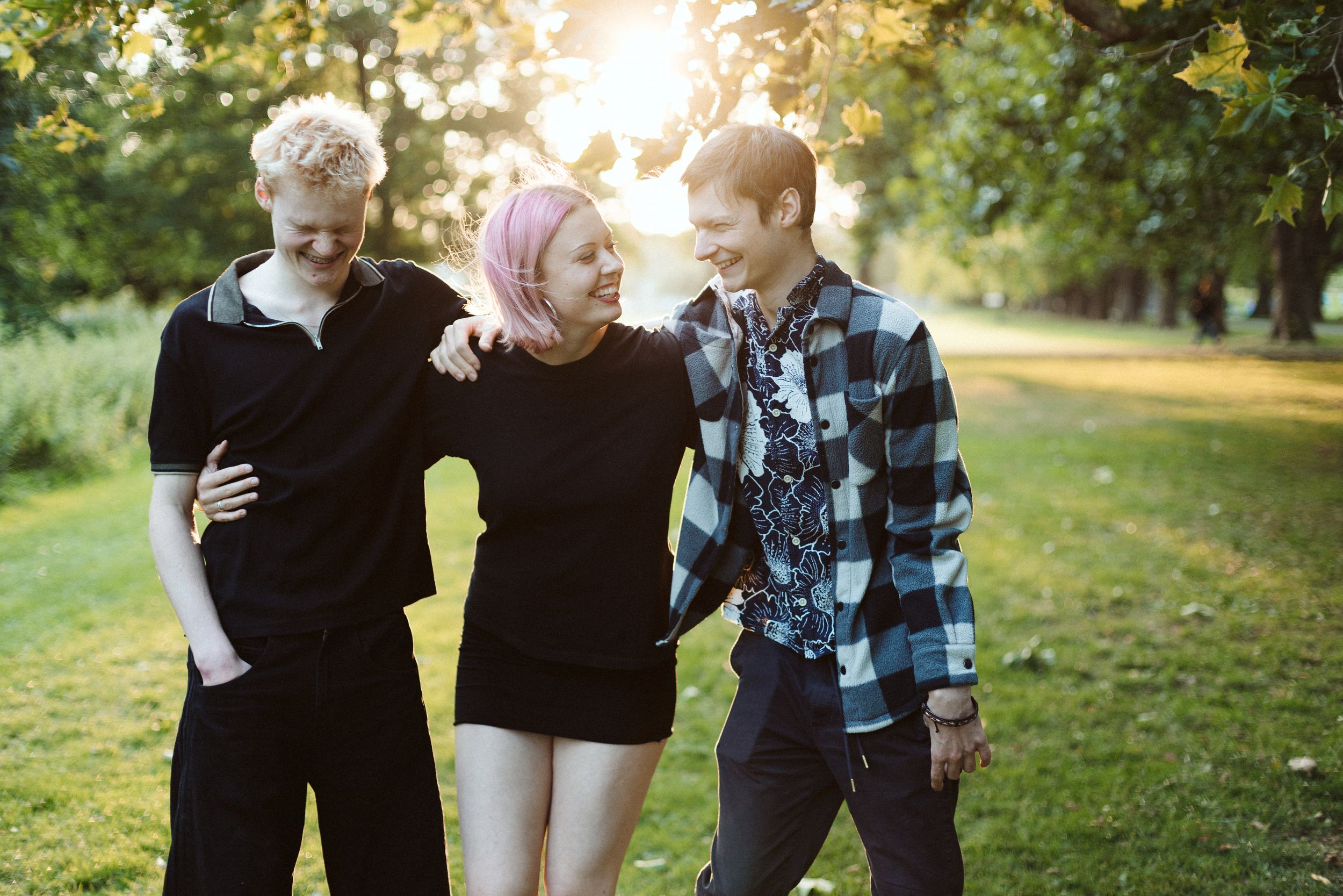 Three smiling friends hugging and walking together in a sunny green park, one with pink hair wearing a black dress.