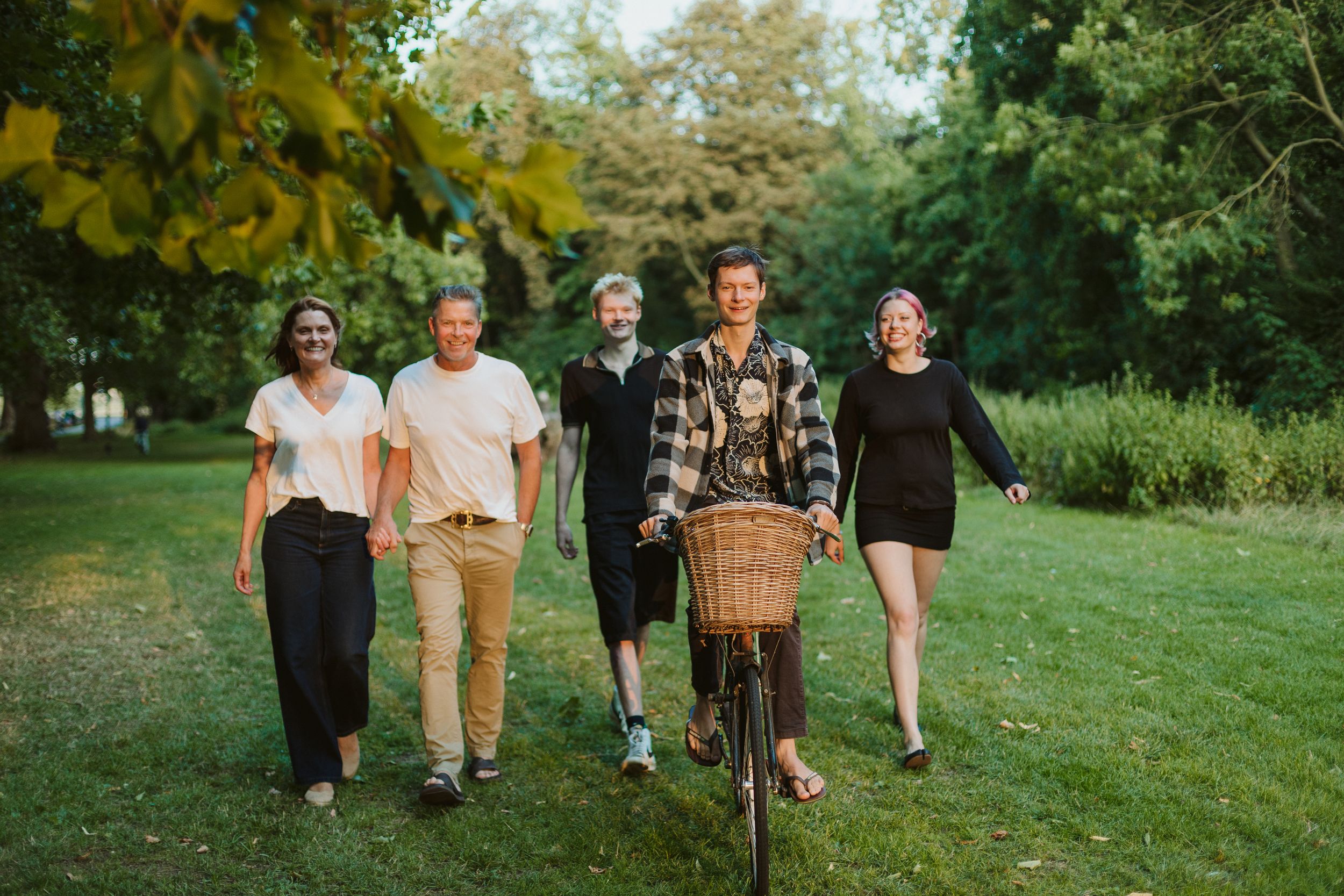 Five friends walk together in a lush green park, one person pushing a vintage bicycle with a wicker basket.