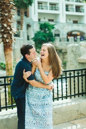 Happy couple embracing and laughing outdoors near a decorative railing with a resort building and palm tree in the background.