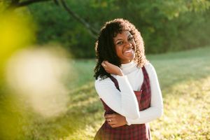 A joyful person in a white turtleneck and burgundy jumper dress stands in golden sunlight.