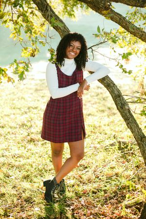 A person in a plaid dress and white sweater poses playfully by a blooming tree.