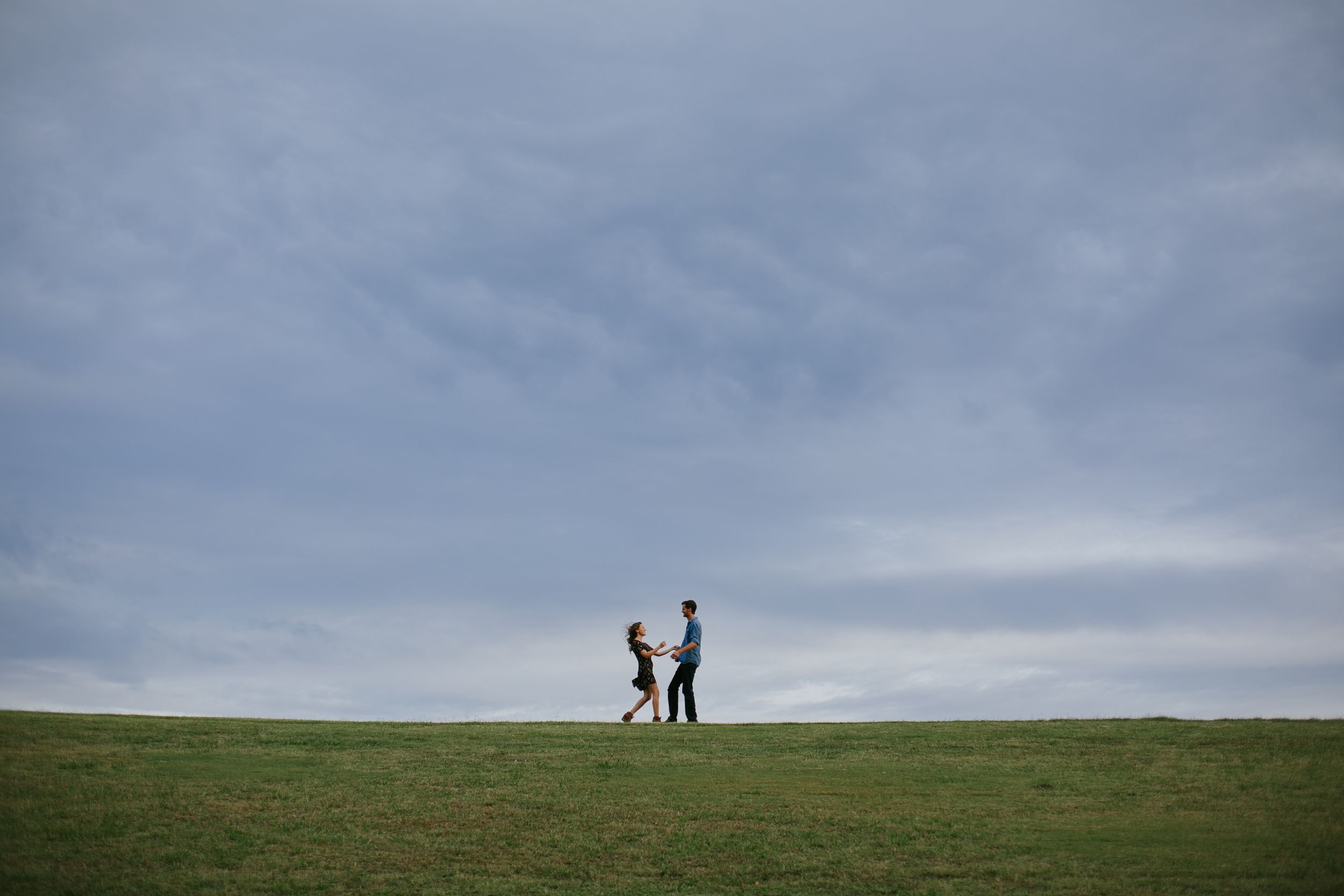 Two figures walking together on grassy hilltop against cloudy sky.