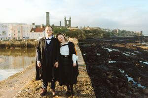 Two people stand by the coast with historic buildings and church towers visible in the background.