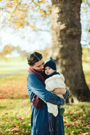 A parent in a blue coat embraces a bundled-up baby on an autumn day under a large tree.