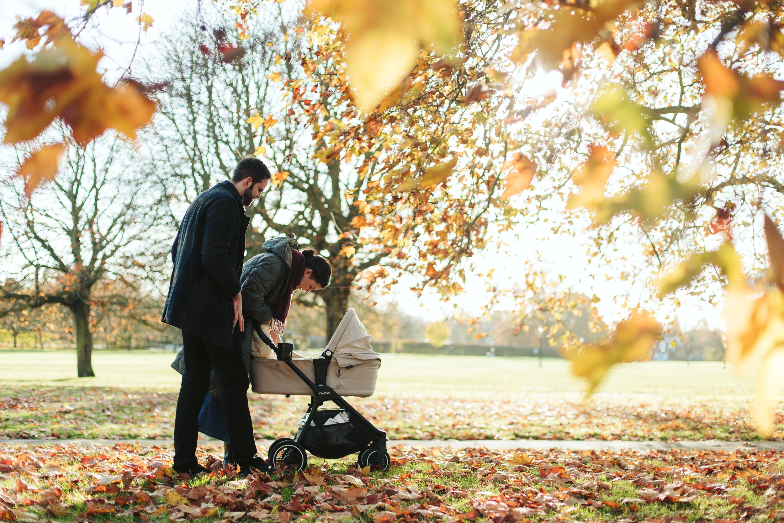 Couple walking with stroller through fallen autumn leaves in a park at sunset.