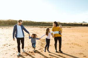 A family of four walks hand in hand along a sunny beach at sunset.