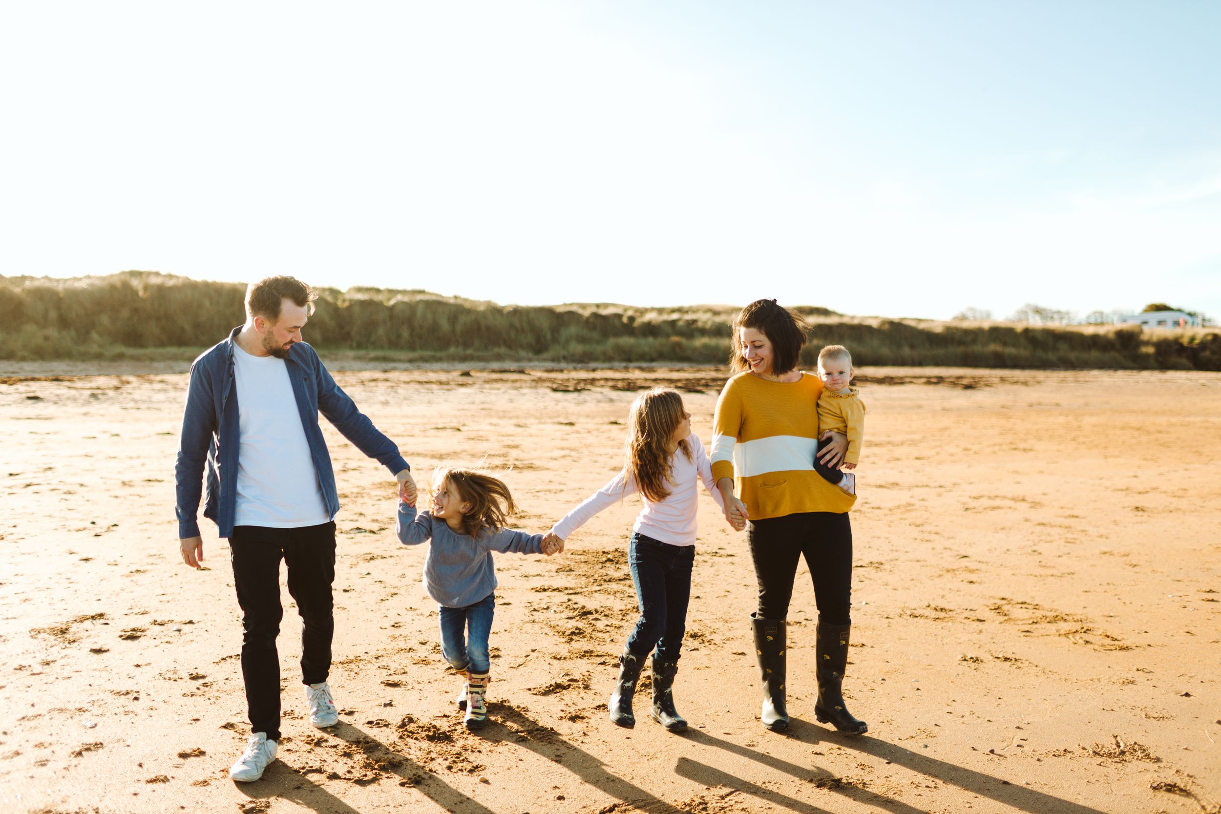 A family of four walks hand in hand along a sunny beach at sunset.