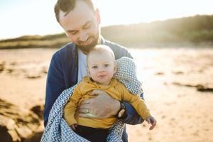 A parent holds a baby wearing yellow on a sunny beach.