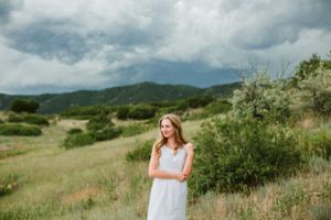 Person in white dress stands in a meadow with cloudy mountain landscape behind.