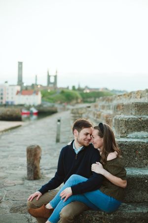 A couple shares an intimate moment on stone steps near a coastal harbor.