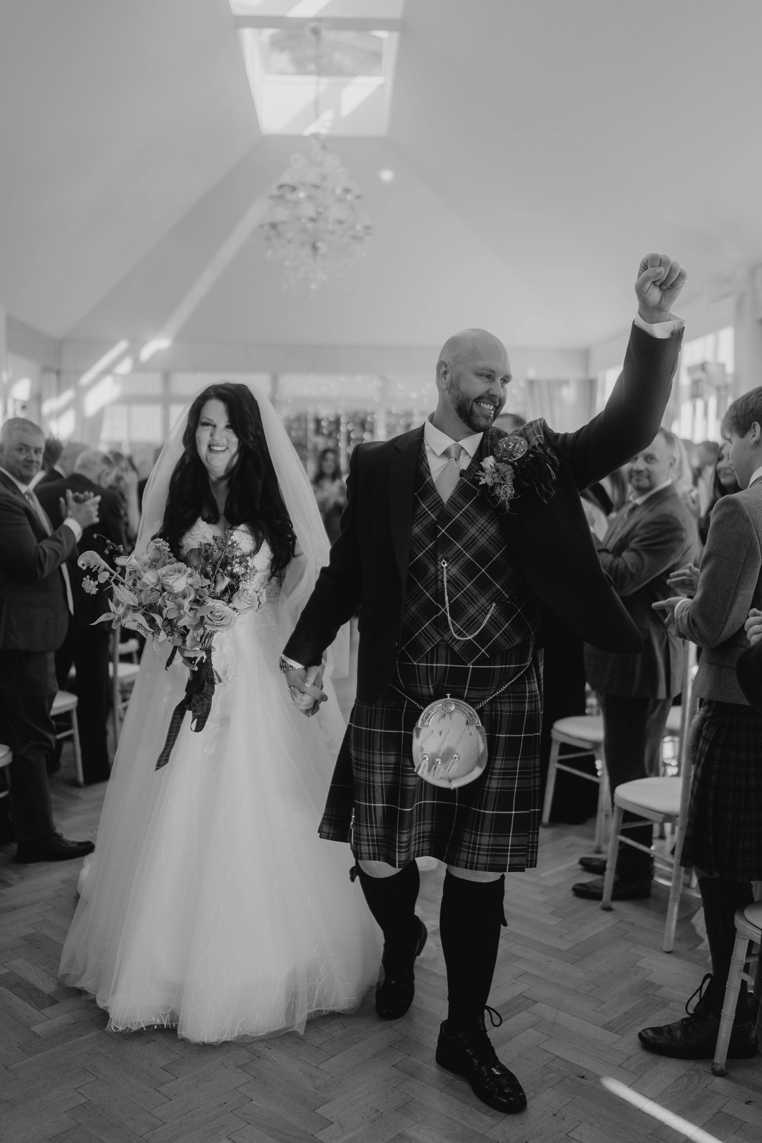 A celebration in a bright venue with traditional Scottish wedding attire and raised arms in triumph.