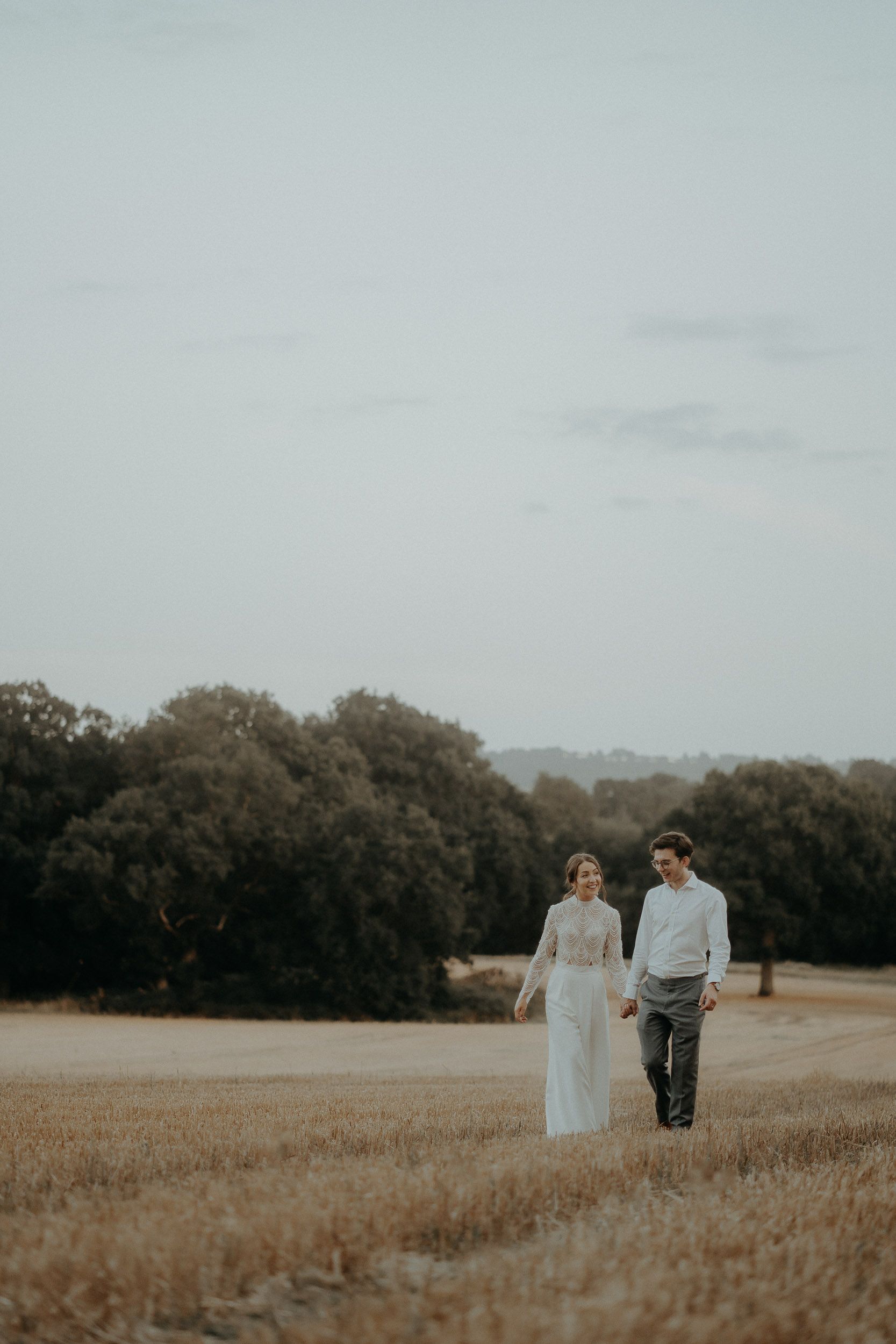 Couple walking hand in hand through golden field with treeline in background.