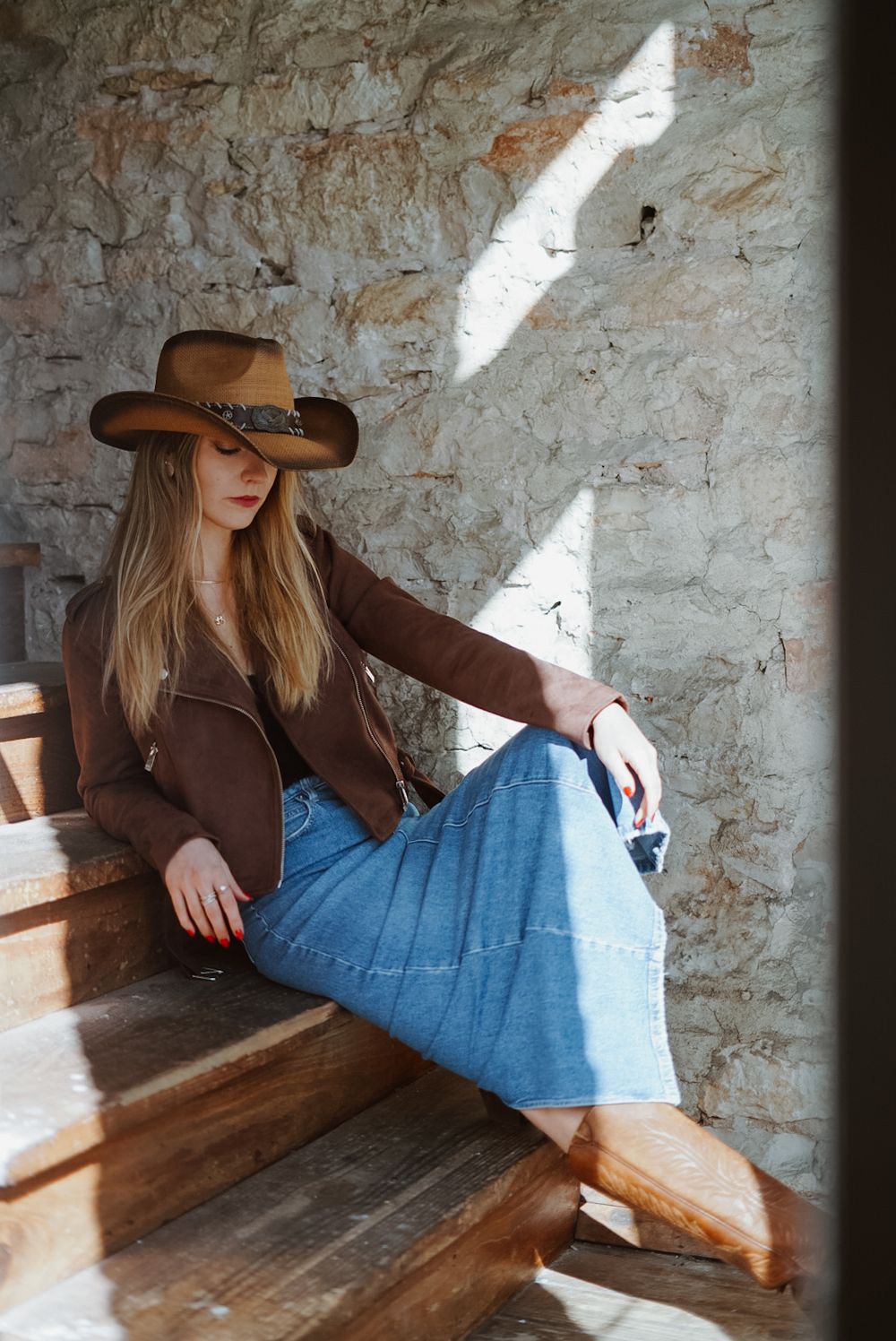 Western-style portrait with brown jacket and cowboy hat against rustic wall with dramatic lighting.