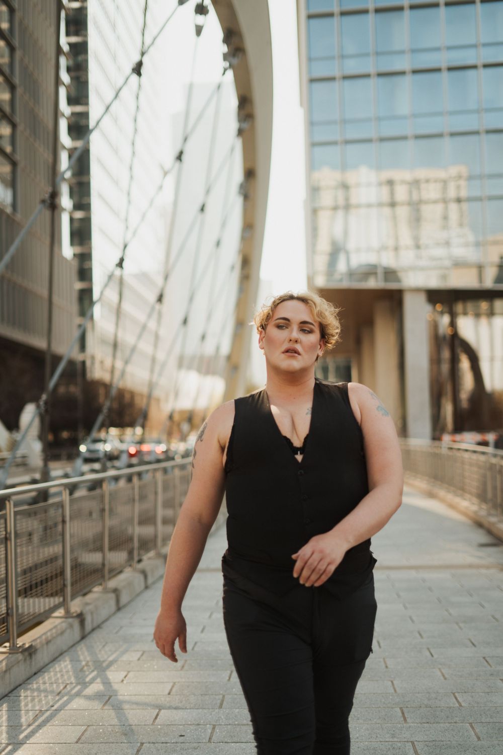 Person in all black outfit poses confidently on urban walkway with modern architecture and cables in background.