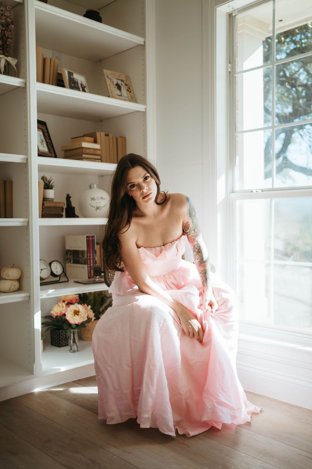 Person in pink gown sits elegantly by window with white built-in shelving.