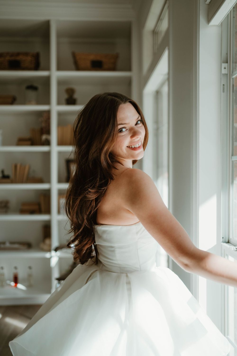 Person in white dress spins joyfully by window and built-in shelves.