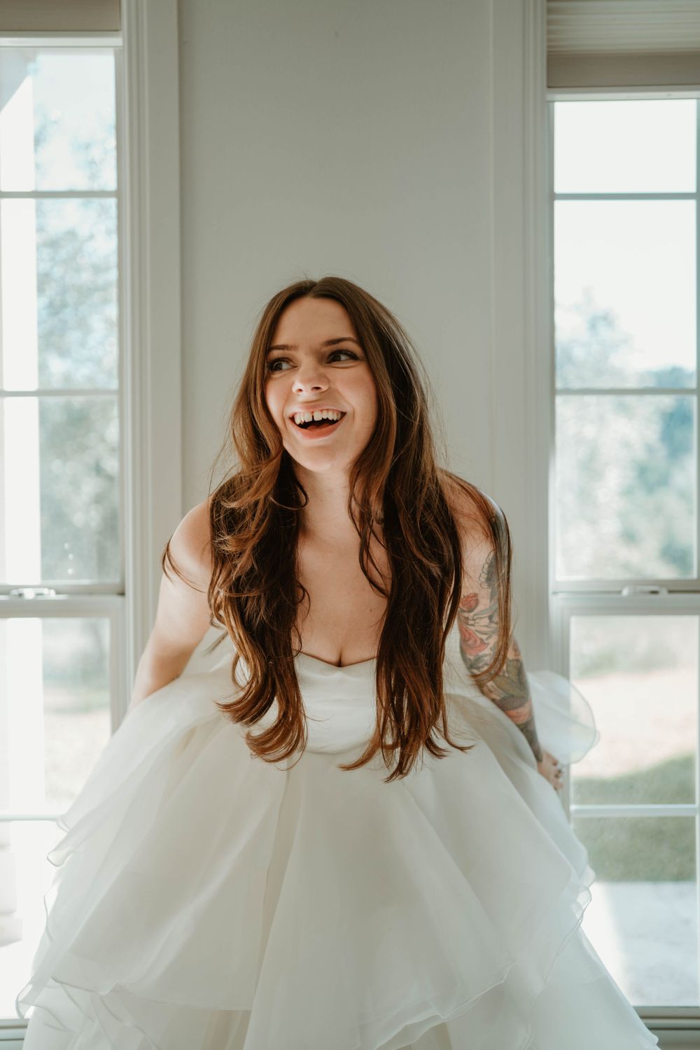 A joyful bride in a white wedding dress laughs while posing near bright windows.