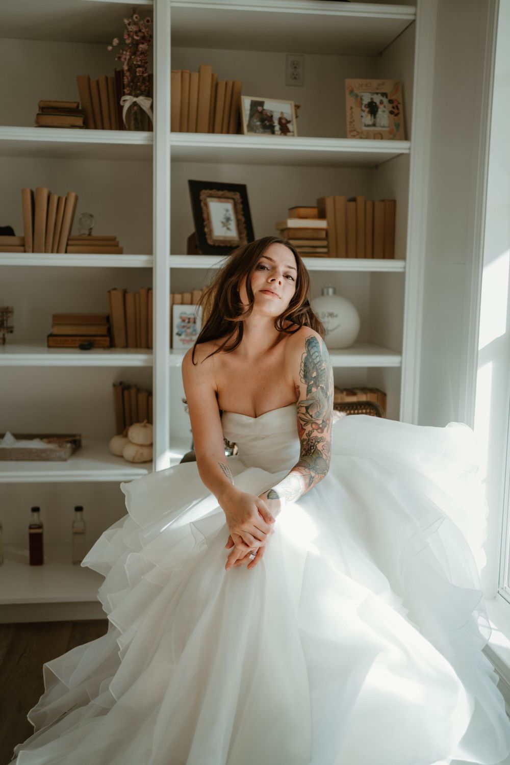 Bride in strapless ballgown poses by bookshelf with natural window light.