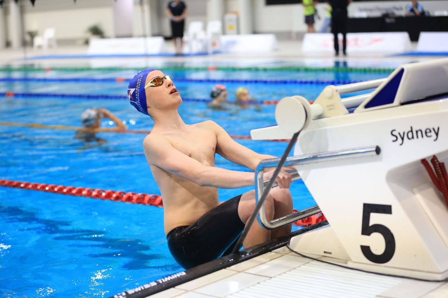 Swimmer in blue cap resting at pool edge next to lane marker number 5.