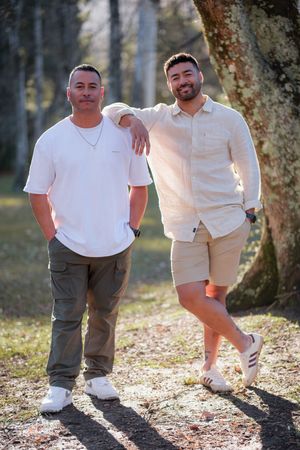 Two people posing together in a wooded outdoor setting wearing casual white and beige clothing.