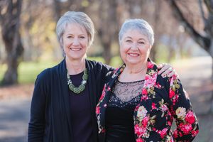 Two women wearing floral and black outfits smile while standing together outdoors.
