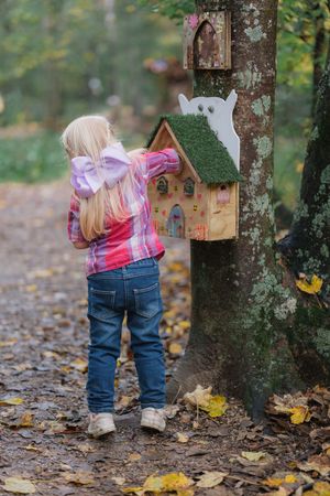 Small child in jeans and plaid shirt looking at wooden birdhouse mounted on tree.