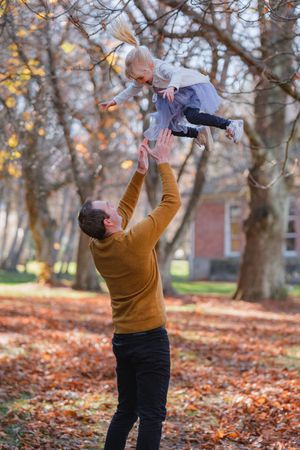 Playful moment captured in autumn leaves as person lifts child in air against fall foliage background.