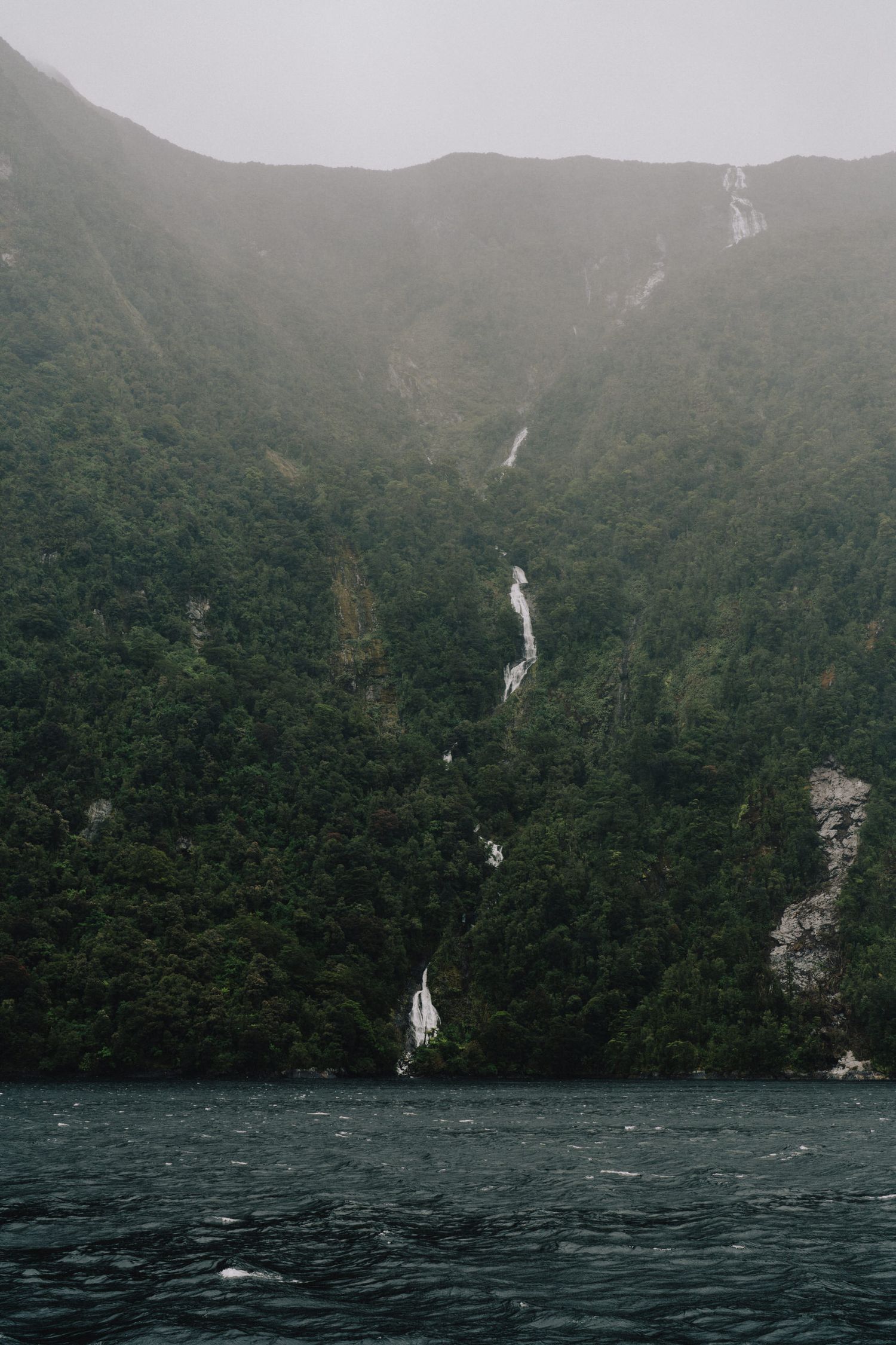 Tall waterfall streaming down a forested mountain cliff into dark waters below.