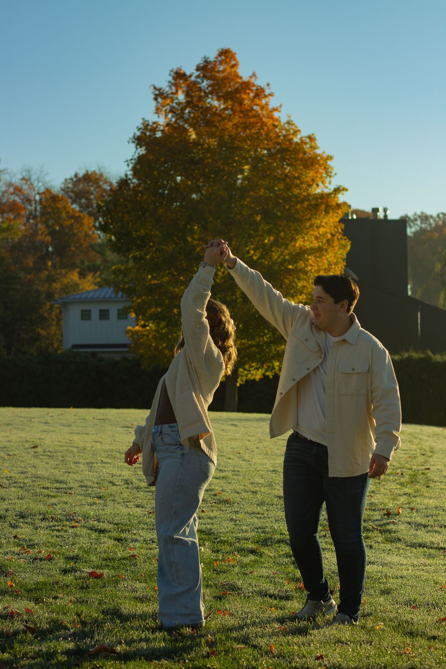 A couple dances together in a grassy field during golden hour with autumn trees and a house in the background during autumn photos.