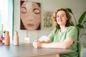 A person in a green shirt sits at a table with pink bottles and artwork displayed on the wall behind them.