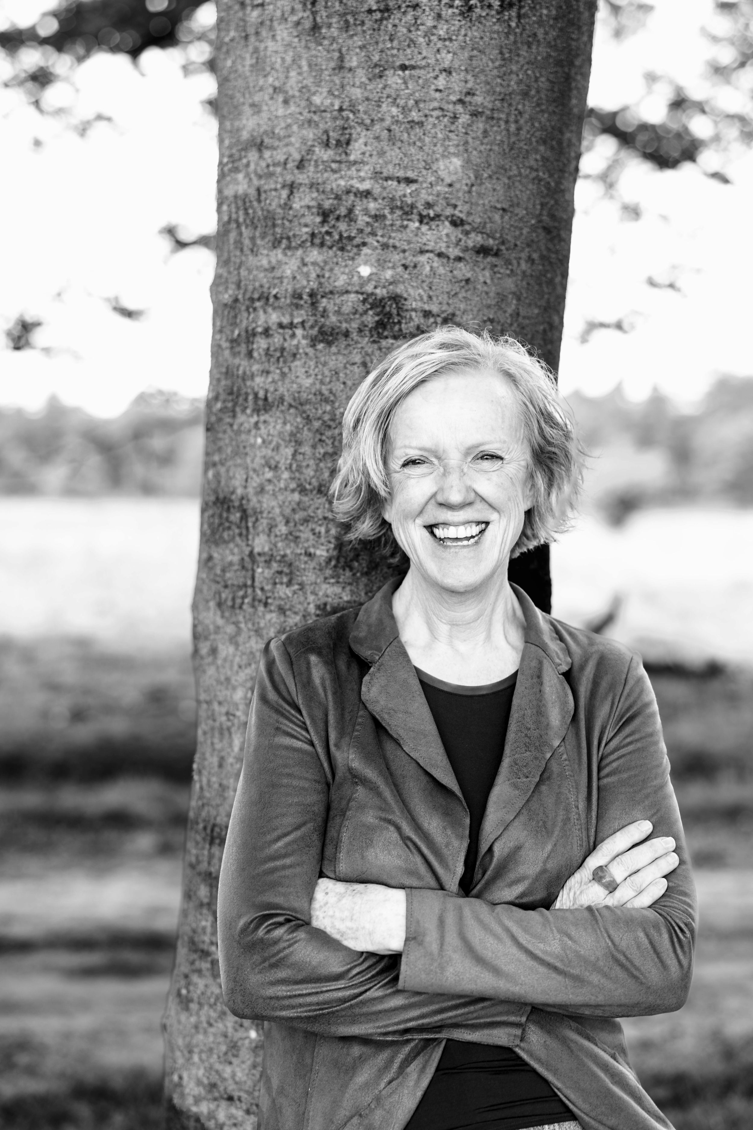 A person stands against a tree trunk wearing a jacket and smiling in black and white.