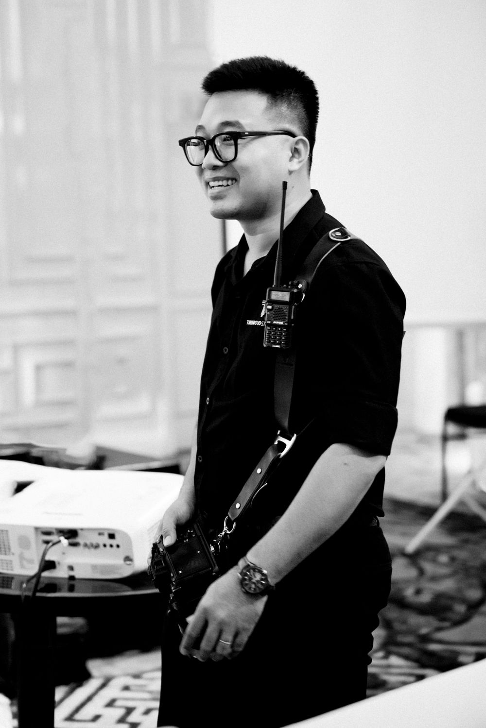 Smiling Asian man in black uniform with glasses and radio, standing in elegant indoor venue, black and white photo.
